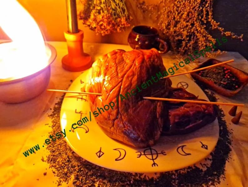 May include: A raw, reddish-brown heart impaled with wooden skewers, presented on a yellow plate adorned with crescent moon and symbol designs. The plate is surrounded by dried herbs and incense cones, with a candle and cup in the background.