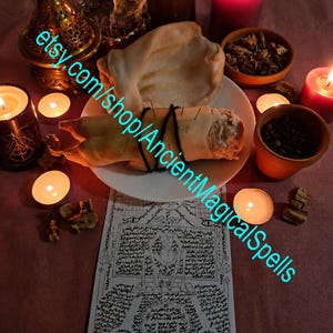 May include: A white plate holds a light-colored, textured object bound with black string and pierced by thin metal pins. The plate sits on a paper document covered in intricate black ink illustrations and text, surrounded by lit candles and bowls.