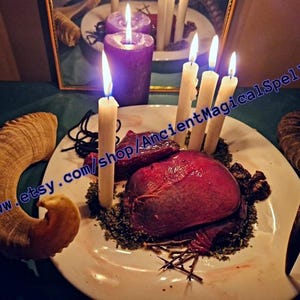 May include: A ritualistic arrangement featuring a white plate with a large, red object surrounded by greenery and several lit white candles. Two curved horns flank the plate. A purple candle and a mirror are in the background.