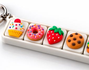 May include: A rectangular white keyring featuring five miniature food-themed charms arranged in a row. The charms include a cupcake with a cherry, a pink frosted doughnut, a red strawberry, a chocolate chip cookie, and a small hamburger.