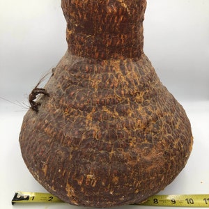 Puede incluir: Un jarrón grande, marrón, tejido a mano, con forma de calabaza y superficie texturizada. El jarrón tiene un cuello estrecho y una base ancha. Tiene un diámetro de aproximadamente 25 cm.