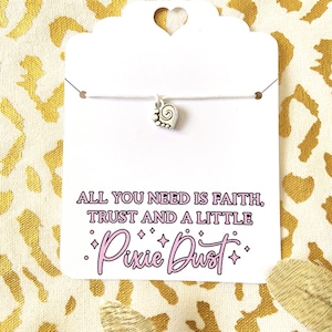 May include: A silver-toned charm shaped like a spiral foot on a thin white string, attached to a white display card. The card features the black and pink text: ALL YOU NEED IS FAITH, TRUST AND A LITTLE Pixie Dust, decorated with small star icons.