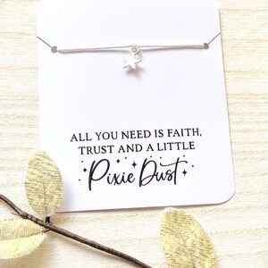 May include: A white card with a heart-shaped cutout, displaying a silver star charm bracelet. The card has the text "ALL YOU NEED IS FAITH, TRUST AND A LITTLE Pixie Dust" in black. A gold leaf branch is in the foreground.