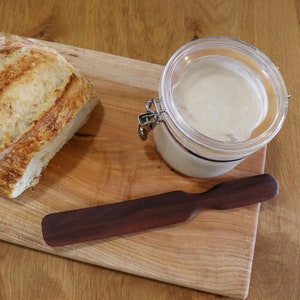 May include: A wooden cutting board with a loaf of sourdough bread, a glass jar of sourdough starter, and a wooden butter knife.
