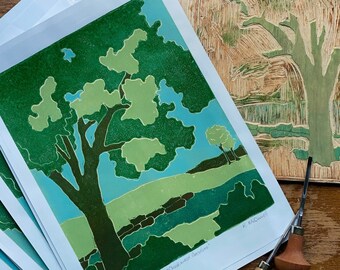 May include: A stack of prints of a green tree with a blue sky and green grass. The print is titled "Cultivated Garden" and signed by the artist, K. McMannis. The print is next to a woodblock carving of a tree.