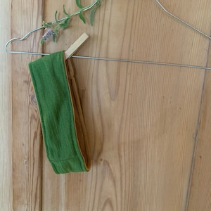 Puede incluir: Una diadema verde y marrón colgada de una percha de alambre. La diadema está hecha de un tejido suave y elástico.