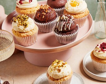 May include: A pink cake stand with a variety of cupcakes. The cupcakes are topped with different frostings and decorations, including chocolate, white chocolate, pistachio, and raspberry.