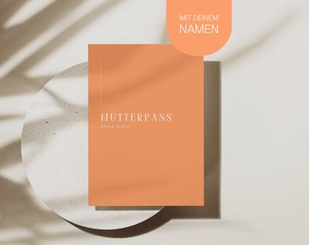 May include: A peach-colored card with the text "MUTTERPASS" and "KARLA GLUCK" in white. The card is on a light brown surface with a shadow of a plant.