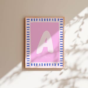 Könnte beinhalten: Ein gerahmter Kunstdruck mit einem großen weißen Buchstaben "A" auf rosa Hintergrund. Der Rahmen hat einen blauen gepunkteten Rand. Der Druck wird an einer beigen Wand mit Schatten angezeigt.