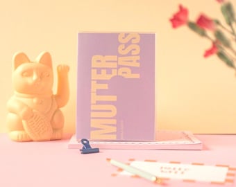 May include: A purple card with the text "MUTTER PASS" in yellow letters. The card is sitting on a pink surface with a yellow cat figurine and a blue binder clip.
