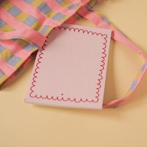 Könnte beinhalten: Eine rosa-gelb gestreifte Tragetasche mit einem blauen Reißverschluss. Die Tasche liegt auf einer hellgelben Oberfläche mit einem rosa Notizblock mit rotem, gewelltem Rand.
