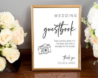 May include: A white sign with a gold frame that says "WEDDING guestbook". There is a black and white illustration of a camera with a heart in the viewfinder. The text below the illustration says "Take a photo, place it in the book and write a message for the couple. Thank you CHLOE & JAMES".