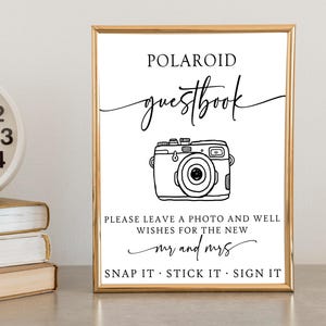 May include: A white sign with a gold frame that says "POLAROID guestbook" in black text. There is a black and white illustration of a camera with the text "PLEASE LEAVE A PHOTO AND WELL WISHES FOR THE NEW mr and mrs SNAP IT STICK IT SIGN IT".