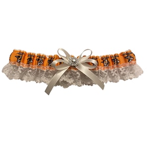 May include: An orange and black garter with a white lace trim and a white satin bow. The garter features a decorative design and a rhinestone embellishment in the center of the bow. The garter is designed for a special occasion.