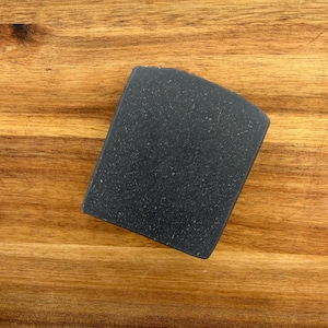 May include: A dark gray, rectangular bar of soap with a rounded top. The soap has a speckled texture and is set against a wooden background.