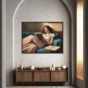 May include: A woman in a white shirt and pink skirt is sitting on a blue couch and reading a book. The couch is in a room with a brown wooden cabinet and a rug.