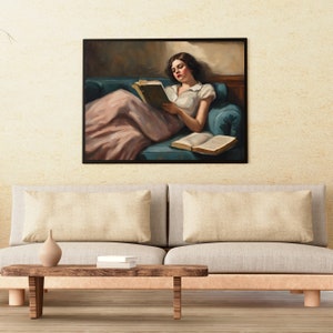 May include: A framed painting of a woman in a white shirt and pink skirt, sitting on a blue couch and reading a book. The painting is hanging on a beige wall.
