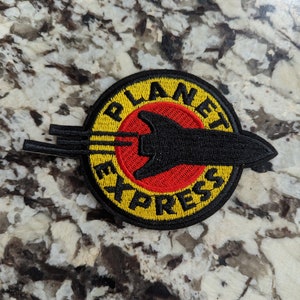 May include: A circular patch with the words "PLANET EXPRESS" in yellow lettering on a black background. The center features a red circle with a black spaceship design. The patch is likely for clothing or accessories.