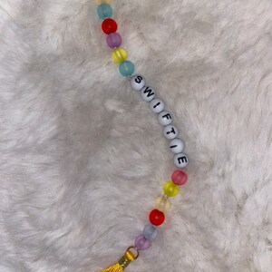 May include: A colourful beaded keychain with a tassel. The beads are translucent and come in shades of yellow, blue, red, and purple. The keychain spells out "SWIFTIE" in white letters.