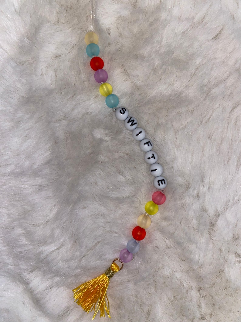 May include: A colourful beaded keychain with a tassel. The beads are translucent and come in shades of yellow, blue, red, and purple. The keychain spells out "SWIFTIE" in white letters.