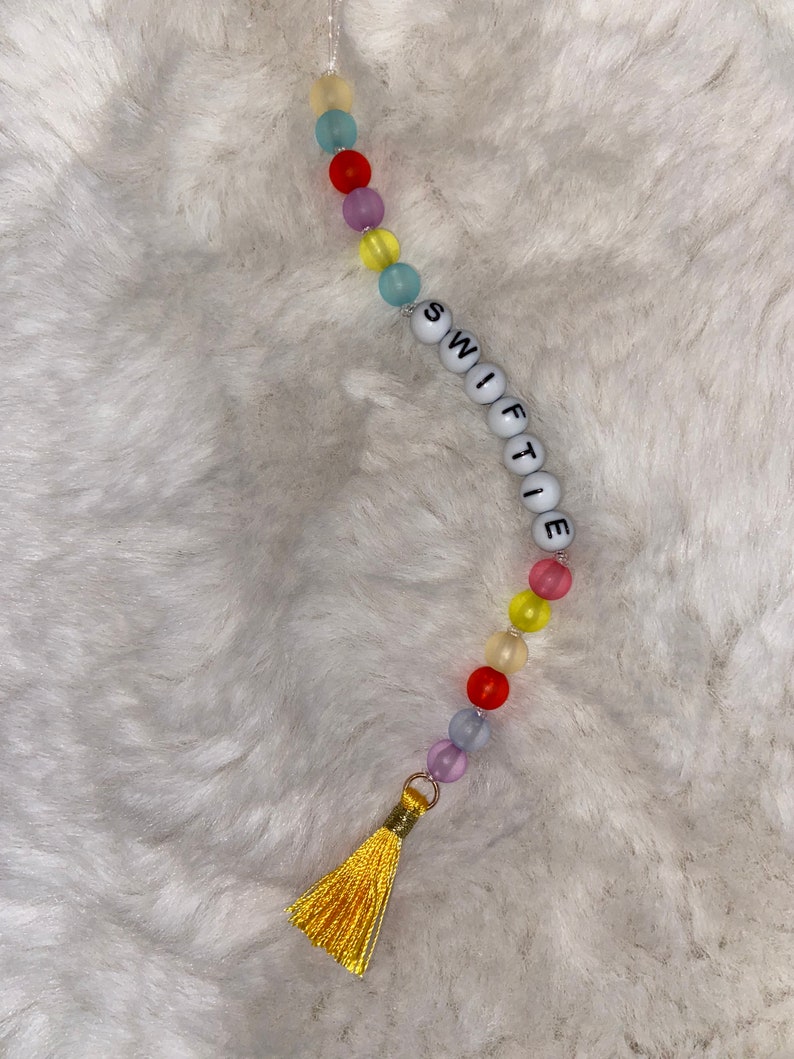May include: A colourful beaded keychain with a yellow tassel. The beads spell out the word "SWIFTIE".