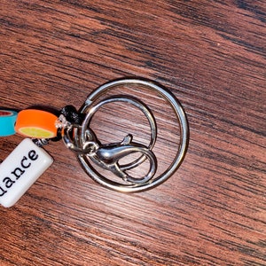 May include: A silver key ring with a small white tag that says "dance" and a colourful charm with orange, blue, and white beads.