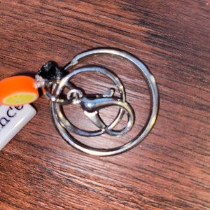 May include: A silver key ring with a round ring and a smaller, attached ring. The keyring has a small orange and white charm attached to it.