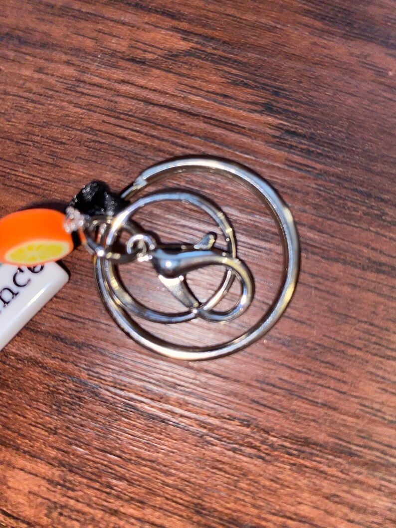 May include: A silver key ring with a round ring and a smaller, attached ring. The keyring has a small orange and white charm attached to it.