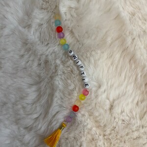 May include: A colourful beaded keychain with the word "SWIFTIE" spelled out in white beads. The keychain has a yellow tassel.