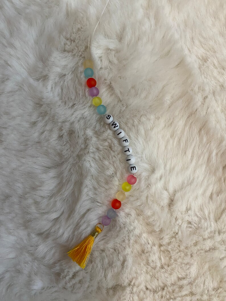 May include: A colourful beaded keychain with the word "SWIFTIE" spelled out in white beads. The keychain has a yellow tassel.