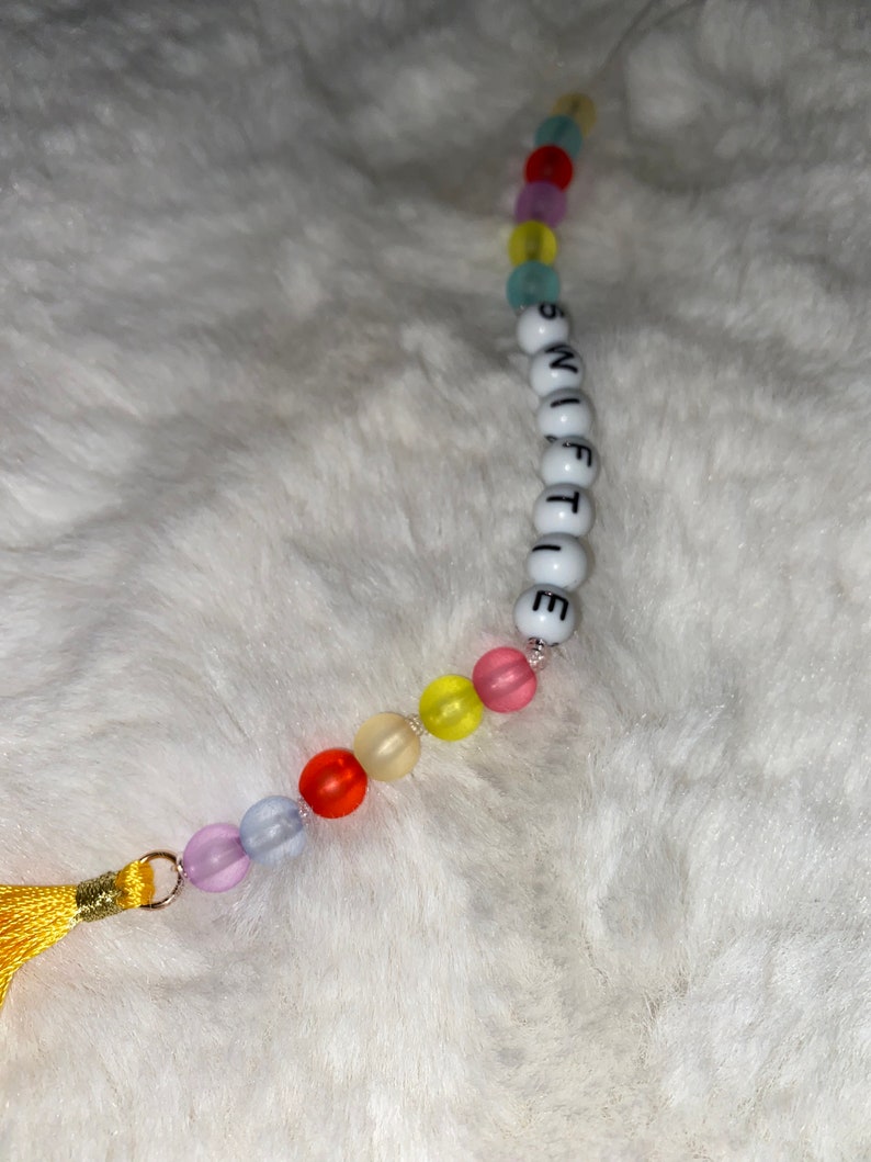 May include: A colourful beaded keychain with a tassel. The beads spell out the word "SWIFTIE".