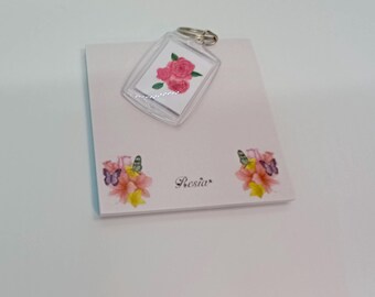 Peut inclure: Un porte-clés rectangulaire transparent avec un anneau argenté présente un motif de roses roses. Le porte-clés repose sur un bloc-notes blanc avec des motifs floraux et papillons et le mot "Presia".