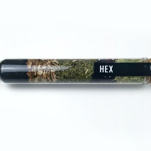 May include: A glass test tube filled with four different types of dried herbs. The tube has a cork stopper and a black label that reads "HEX".