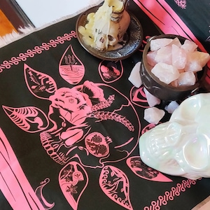 May include: Black fabric with a pink skull and floral design. The fabric is surrounded by a pink border with a repeating pattern. There is a white skull candle and a bowl of pink crystals on the fabric.