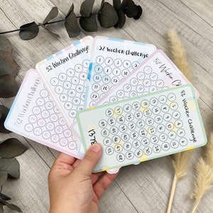 Könnte beinhalten: Vier farbige Karten mit dem Text "52 Wochen Challenge" und einem Raster mit Kreisen, die von 1 bis 52 nummeriert sind. Die Karten sind fächerförmig angeordnet und werden in einer Hand gehalten.