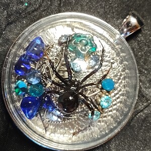 Puede incluir: Un colgante de resina transparente con una araña negra, una telaraña plateada y cuentas de vidrio azules y verdes.