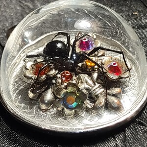 Puede incluir: Una araña negra con piedras preciosas rojas, moradas y amarillas está encerrada en una cúpula de vidrio redonda plateada. La cúpula se asienta sobre una base plateada con un diseño decorativo.