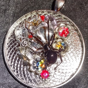 Puede incluir: Un colgante de plata con una araña negra y flores y cuentas de colores. La araña está en el centro del colgante y está rodeada de flores y cuentas.