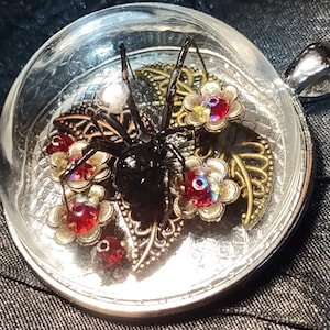 Puede incluir: Un colgante con una araña negra dentro de una cúpula de cristal transparente. La araña está rodeada de filigrana dorada y adornos en forma de flor rojos e iridiscentes. El colgante tiene una fianza plateada.