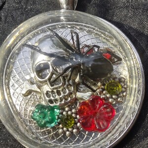 Puede incluir: Un colgante de calavera de plata con una araña negra y flores rojas, verdes y amarillas encapsuladas en resina.