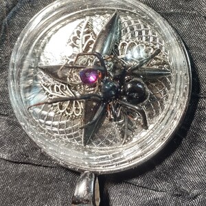 Puede incluir: Un colgante de plata con una araña negra y una gema púrpura en una cúpula de resina transparente.