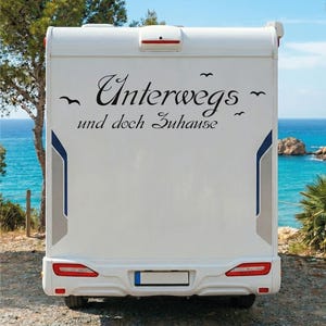 Könnte beinhalten: Ein weißes Wohnmobil mit dem deutschen Text "Unterwegs und doch Zuhause" in schwarzer Schrift. Das Fahrzeug hat rote Rücklichter und einen Bereich für das Nummernschild. Der Hintergrund zeigt blaues Meer und Himmel.