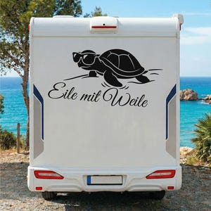 Peut inclure: Un camping-car blanc arbore un autocollant en vinyle noir représentant une tortue portant des lunettes de soleil. Sous la tortue, le texte allemand Eile mit Weile est écrit en écriture cursive noire. Le véhicule a des feux arrière rouges.