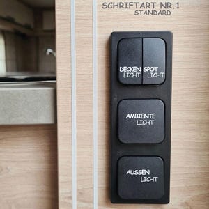 Könnte beinhalten: Ein schwarzes, rechteckiges Lichtschalter-Panel auf einer hellen Holzoberfläche. Das Panel hat vier Tasten mit weißer Beschriftung: Decken Licht, Spot Licht, Ambiente Licht und Aussen Licht. Darüber steht der Text Schriftart Nr. 1 Standard.