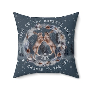 Peut inclure: Un coussin bleu avec un motif hivernal représentant deux lièvres, une couronne de branches de pin et le texte "Même dans la nuit la plus sombre, nous nous réveillons au soleil".