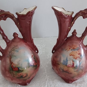 May include: Pair of maroon ceramic vases with ornate handles and a landscape scene painted on the front. The vases have a bulbous base and a flared top with gold trim. The landscape features figures and trees.