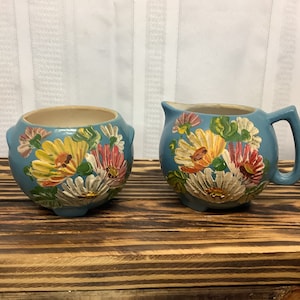 May include: A light blue ceramic creamer and sugar bowl set. The set is decorated with colorful, raised floral designs in yellow, pink, and white. The creamer has a handle and spout.