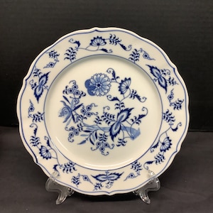 May include: White ceramic plate with a scalloped edge and a blue floral pattern. The plate features a central floral design and a repeating pattern around the rim. The plate is displayed on a clear stand.