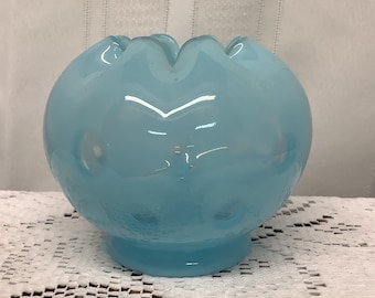 May include: A light blue glass bowl with a scalloped rim and a round base. The bowl has a smooth, glossy finish and is sitting on a white lace tablecloth. The background is a white striped fabric.
