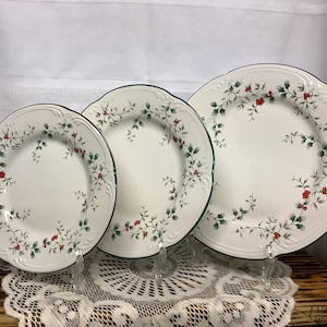 May include: Three white ceramic plates with a green border and a holly berry design. The plates are displayed on clear stands. The plates are different sizes, with the largest plate on the right.